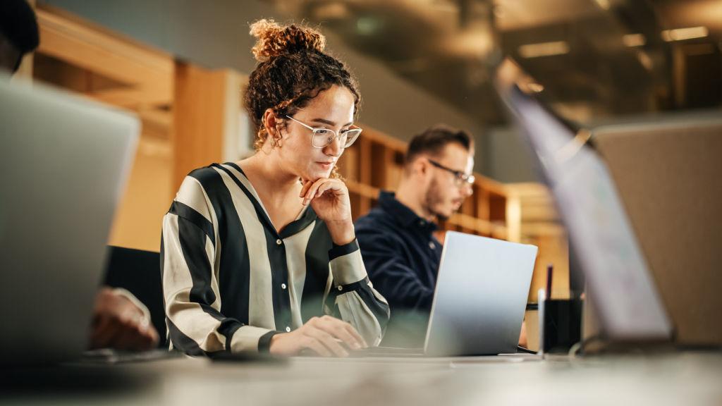 Portrait of Enthusiastic Young Woman Working on Computer in a Modern Bright Office. Confident Human Resources Agent Smiling Happily While Collaborating Online with Colleagues.